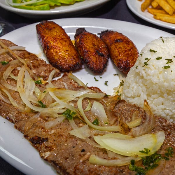 Cuban steak with onions, rice, and fried plantains on a white plate at Rumba Cuban Cafe.