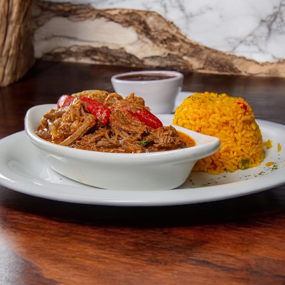 Braised shredded beef topped with red peppers, served with yellow rice and dipping sauce on a white platter.