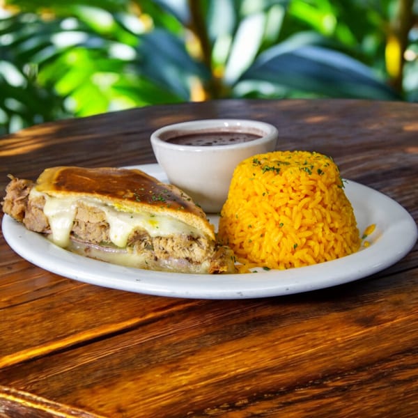 Cuban sandwich with melted cheese, yellow rice, and a side of black beans on a wooden table.