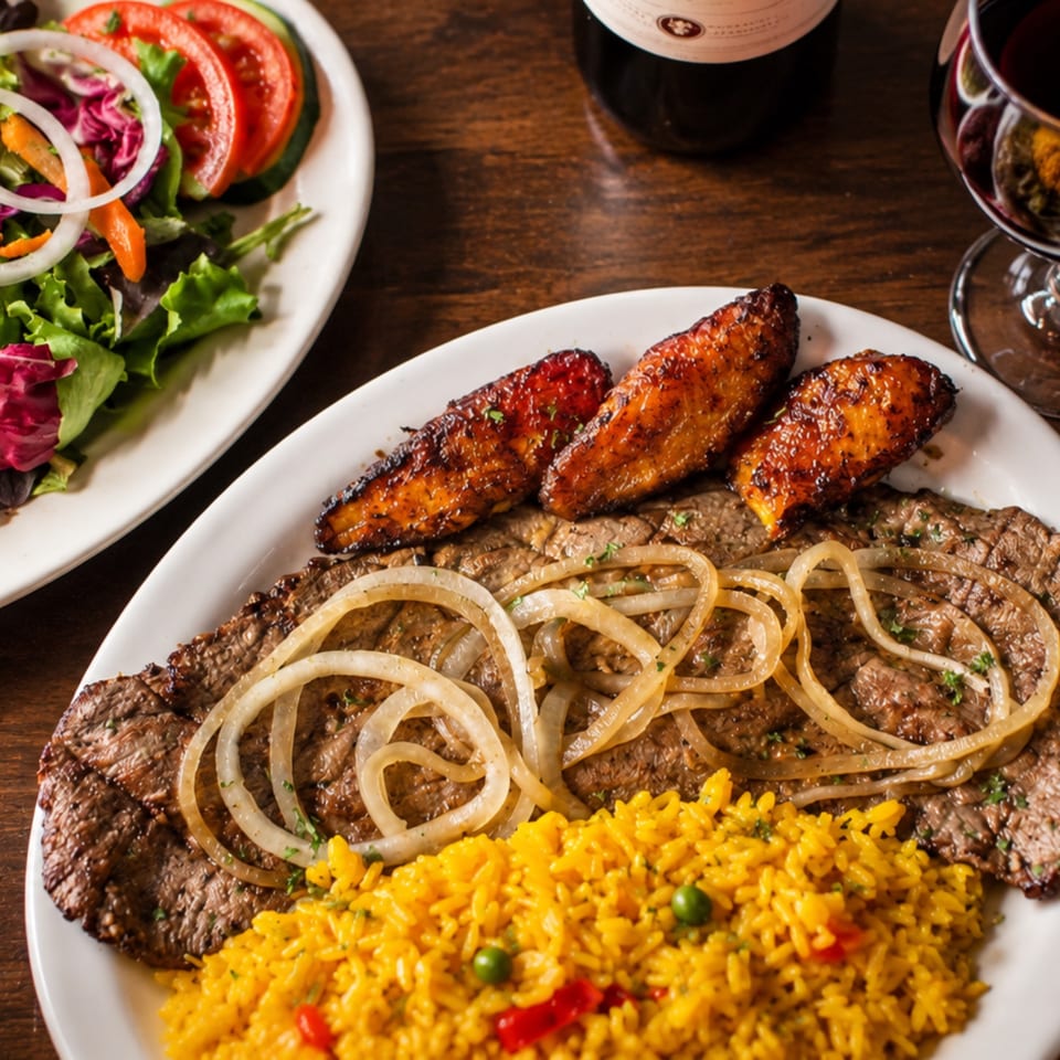 Grilled steak topped with onions, glazed carrots, and yellow rice served beside a fresh garden salad.