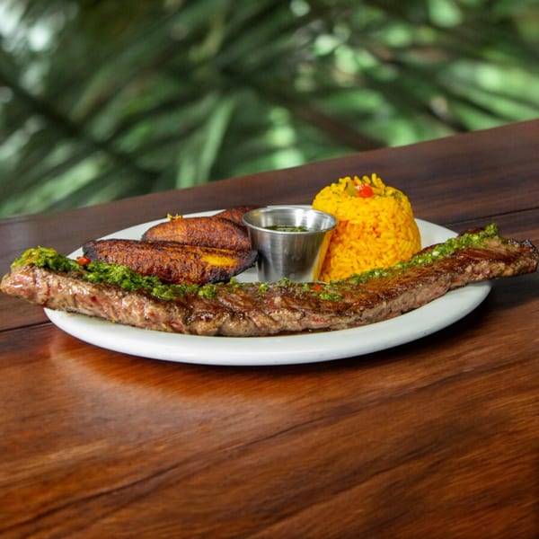 Cuban skirt steak with green sauce, yellow rice, fried plantains, and dipping sauce on a white plate.