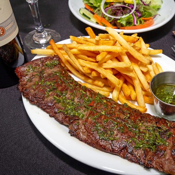 Grilled Cuban steak with chimichurri, a side of fries, and a small salad on a dark table.