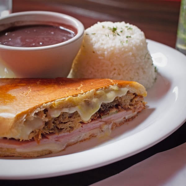 A Rumba Cuban Cafe Cuban sandwich with black beans and white rice on a white plate.