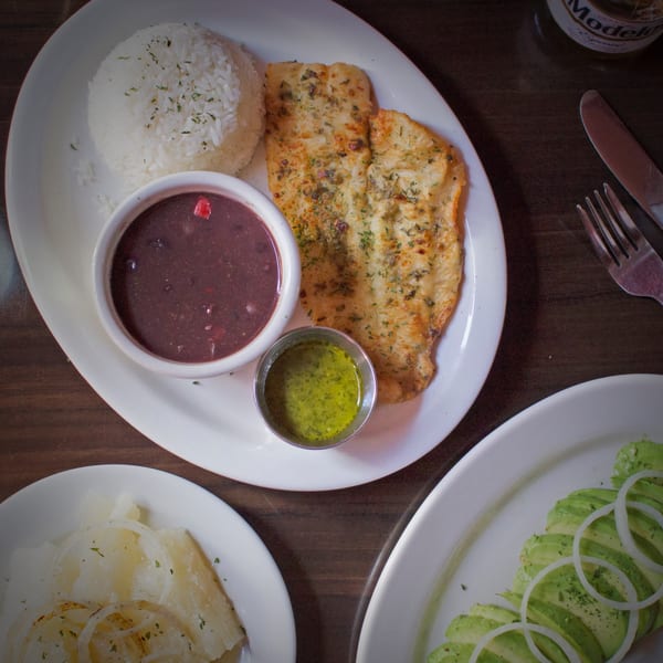 Cuban meal with seasoned fish, white rice, black beans, green sauce, and avocado salad.