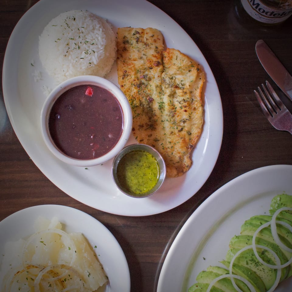 Cuban meal with seasoned fish, white rice, black beans, green sauce, and avocado salad.