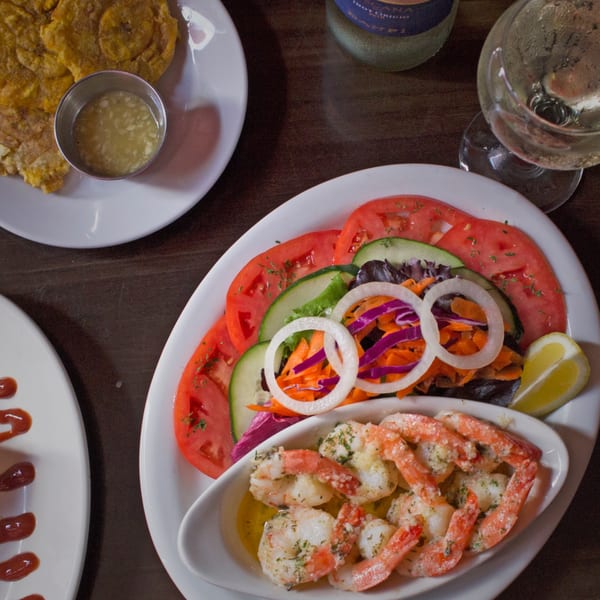 Cuban meal with shrimp, salad, fried plantains, and a glass of white wine on a dark table.