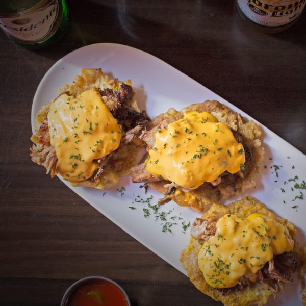Three Cuban tostones topped with shredded pork, melted cheese, and parsley, with beer bottles.