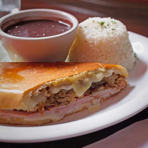 A Cuban sandwich with melted cheese, a side of white rice, and a bowl of black beans.